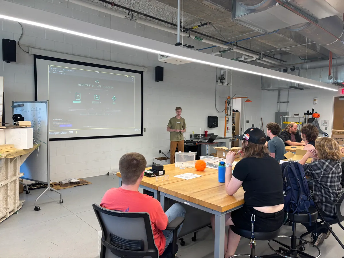 A student presents the Meshtastic Web Flasher on a projector screen to a group seated around a workbench.
