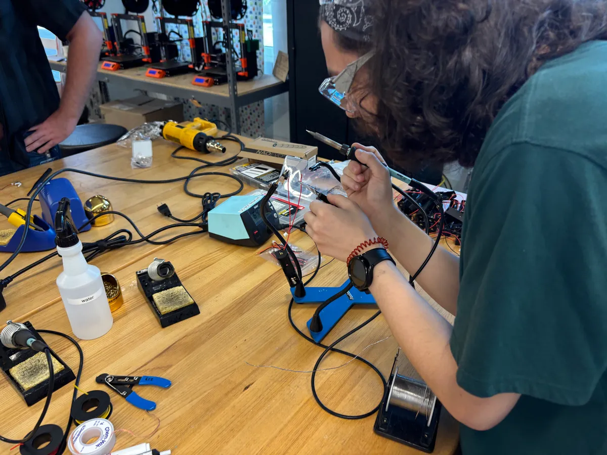 A student in safety glasses solders a battery harness at a bench, with a row of 3D printers running behind them.