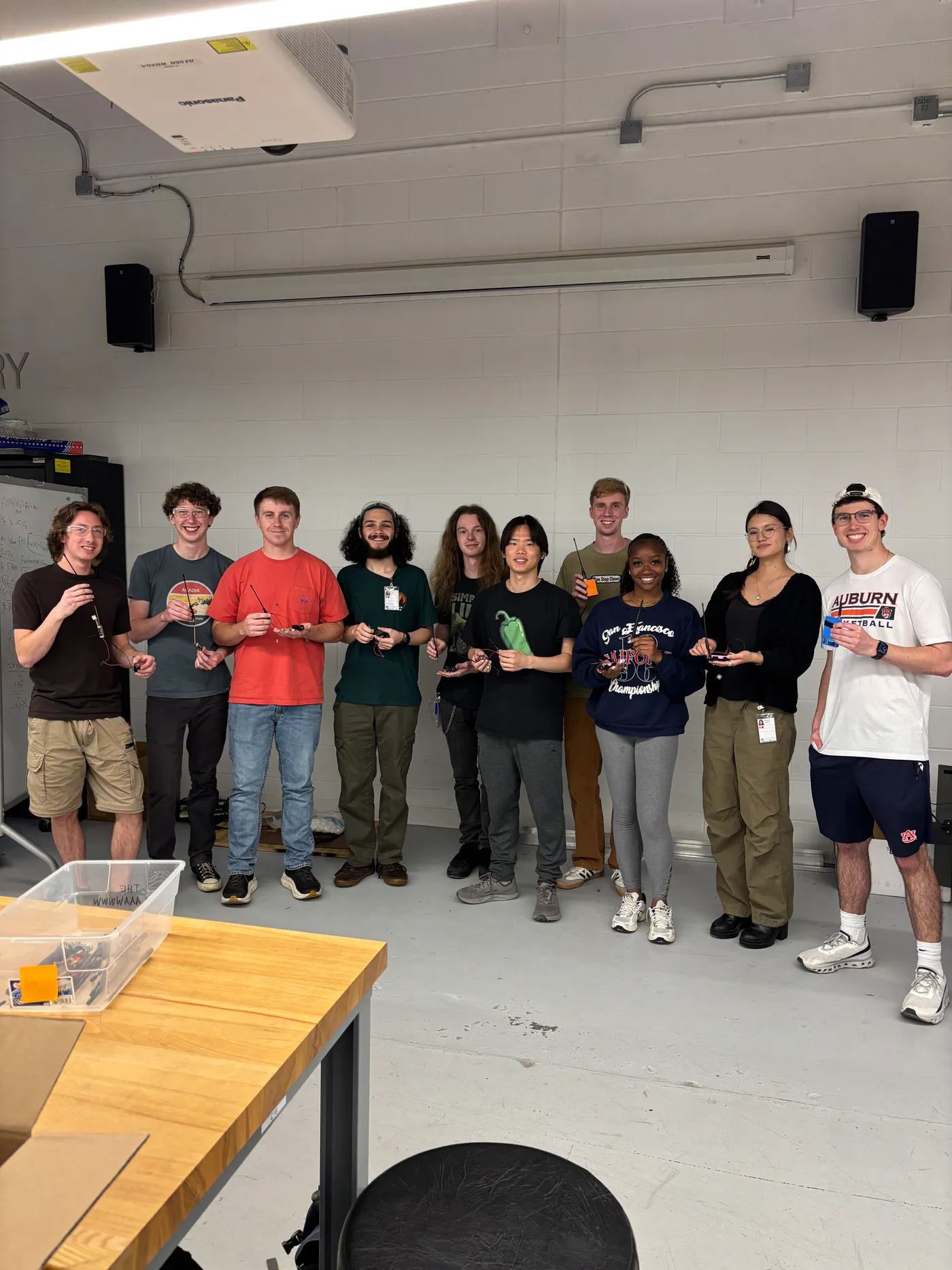 Twelve students standing in a row in the Makerspace, each holding the Meshtastic radio they built.